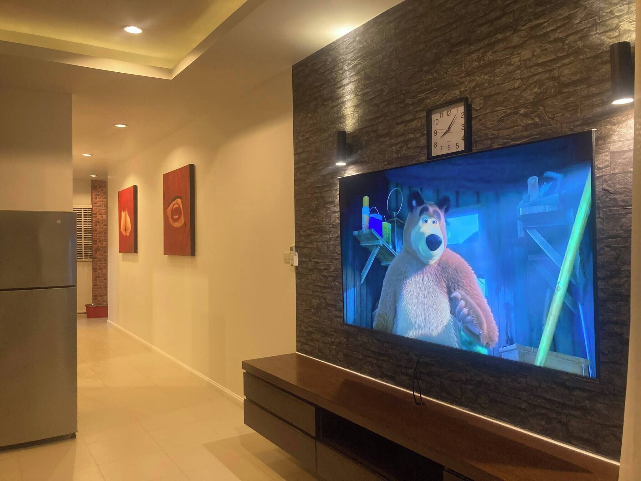 Ein modernes Wohnzimmer mit einem großen Flachbildfernseher an einer Wand mit Steinoptik, einer Holzunterhaltungseinheit darunter und einer Küchenzeile im Hintergrund.