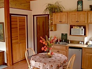 Une table à manger recouverte d’une nappe à motifs floraux, un vase rempli de fleurs et un panier de fruits. On aperçoit également un buffet en bois, avec un four à micro-ondes placé au-dessus.