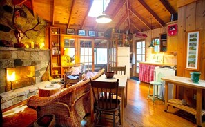 A cozy lodge interior with a stone fireplace, wicker furniture, a dining table, and a kitchen area. - Unique Cabin Vacation Rental for Large Groups on the coast of Lake Michigan near Saugatuck, Michigan (Fennville)