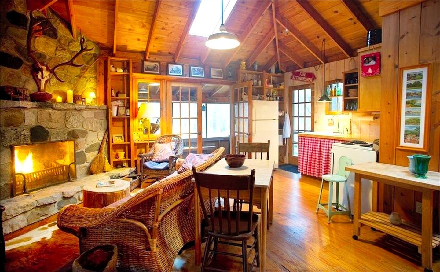 A cozy lodge interior with a stone fireplace, wicker furniture, a dining table, and a kitchen area.