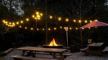 An outdoor area with picnic tables, a fire pit, string lights, and a red umbrella.