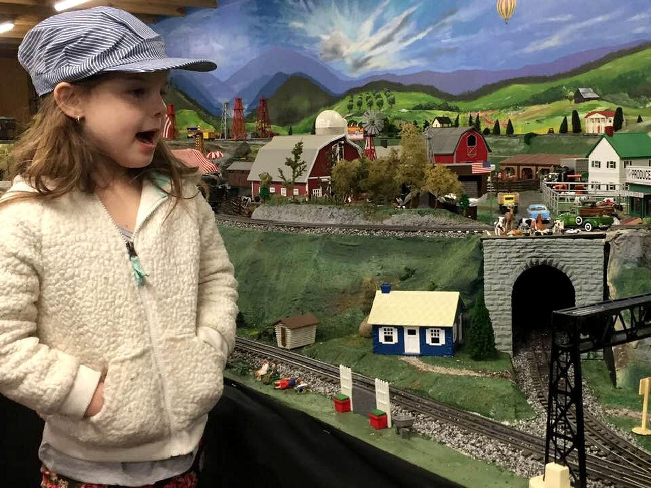 Niño de pie frente a una maqueta de tren con paisaje rural, que incluye un molino, un granero y un puente.