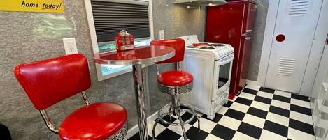 A retro diner interior with a checkered floor, red bar stools, a vintage stove, and a Coca-Cola advertisement.