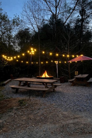 Un espace extérieur aménagé avec des tables de pique-nique, un foyer, des guirlandes lumineuses et un parasol rouge.