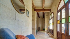 Una habitación con un sillón azul, techo de madera, paredes de piedra y vistas a través de una ventana con contraventanas de madera.