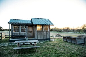 A small wooden cabin with a metal roof, a wooden door, and windows. There is a picnic table in front of the cabin.