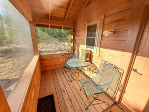 A wooden cabin with a porch, metal chairs, a table, and a window. - Cozy Tiny Cabin - Off-Grid Experience with Sled Dogs Year 'round in Minnesota (Grand Marais)
