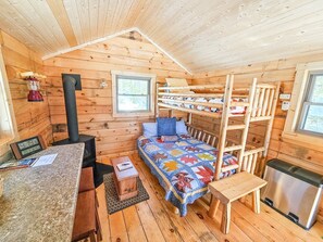 A cozy cabin with a bunk bed, a small kitchen area, a wood-burning stove, and a window. - Cozy Tiny Cabin - Off-Grid Experience with Sled Dogs Year 'round in Minnesota (Grand Marais)