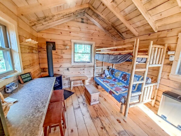A cabin interior with a bunk bed, a wooden desk, a bench, a small table, and a window.