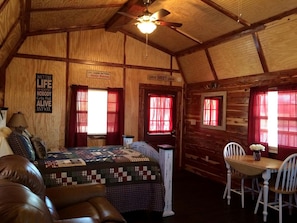 Interior de una cabaña con una cama, una mesa y sillas, y un ventilador de techo.