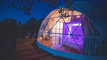 A dome-shaped tent with a transparent structure, a wooden deck, and a purple light inside.
