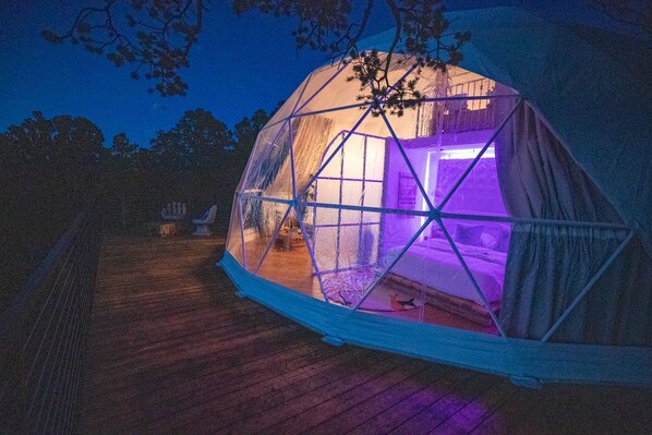 Una tenda a forma di cupola con struttura trasparente, un pontile in legno e una luce viola all'interno.