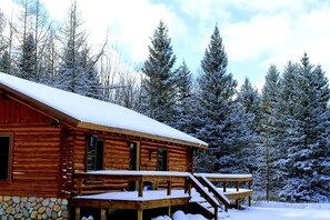 Eine Blockhütte mit Stein Fundament, einer Holzterrasse und einem mit Schnee bedeckten Dach.