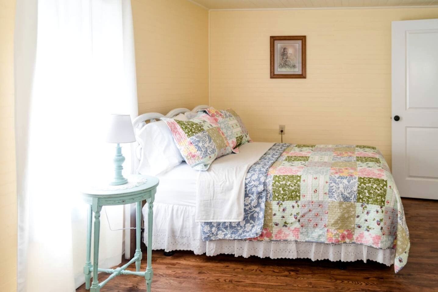 Une chambre à coucher avec un lit, une petite table avec une lampe, une porte et un tableau accroché au mur.