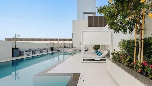 A rooftop pool with lounge chairs, a small seating area with cushions, and a view of a tall building. - Vogue | Luma Park Views JVC Near Circle Mall (Dubai)