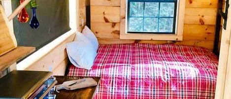 Une petite cabane comprenant un lit, une fenêtre, une étagère avec des objets décoratifs et un bureau avec un ordinateur.