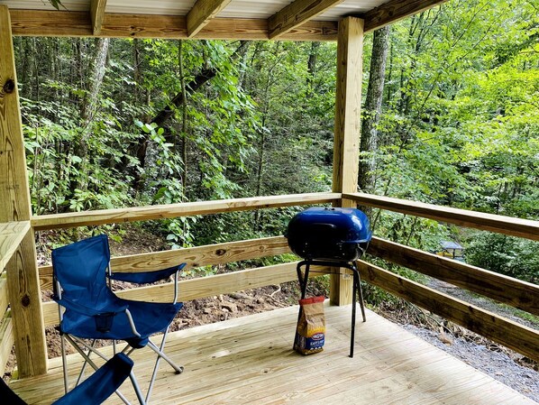 A wooden deck with a barbecue grill, a blue folding chair, and a bag of charcoal. - Cozy and Rustic Cabin Rental in the Smokies for Romantic Getaways in Tennessee (Newport)