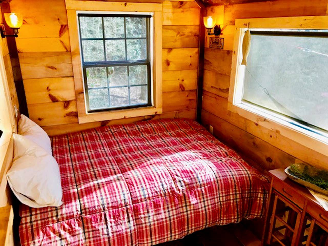 A small cabin room with a plaid bedspread, a window with a view, and a wooden wall.