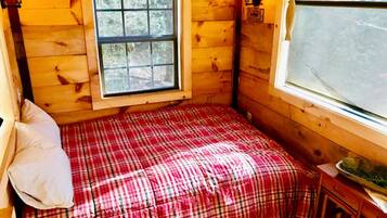 A small cabin room with a plaid bedspread, a window with a view, and a wooden wall.
