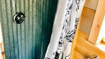 A small, enclosed shower area with a corrugated metal wall, a white shower curtain with a black pattern, and a wooden bench.