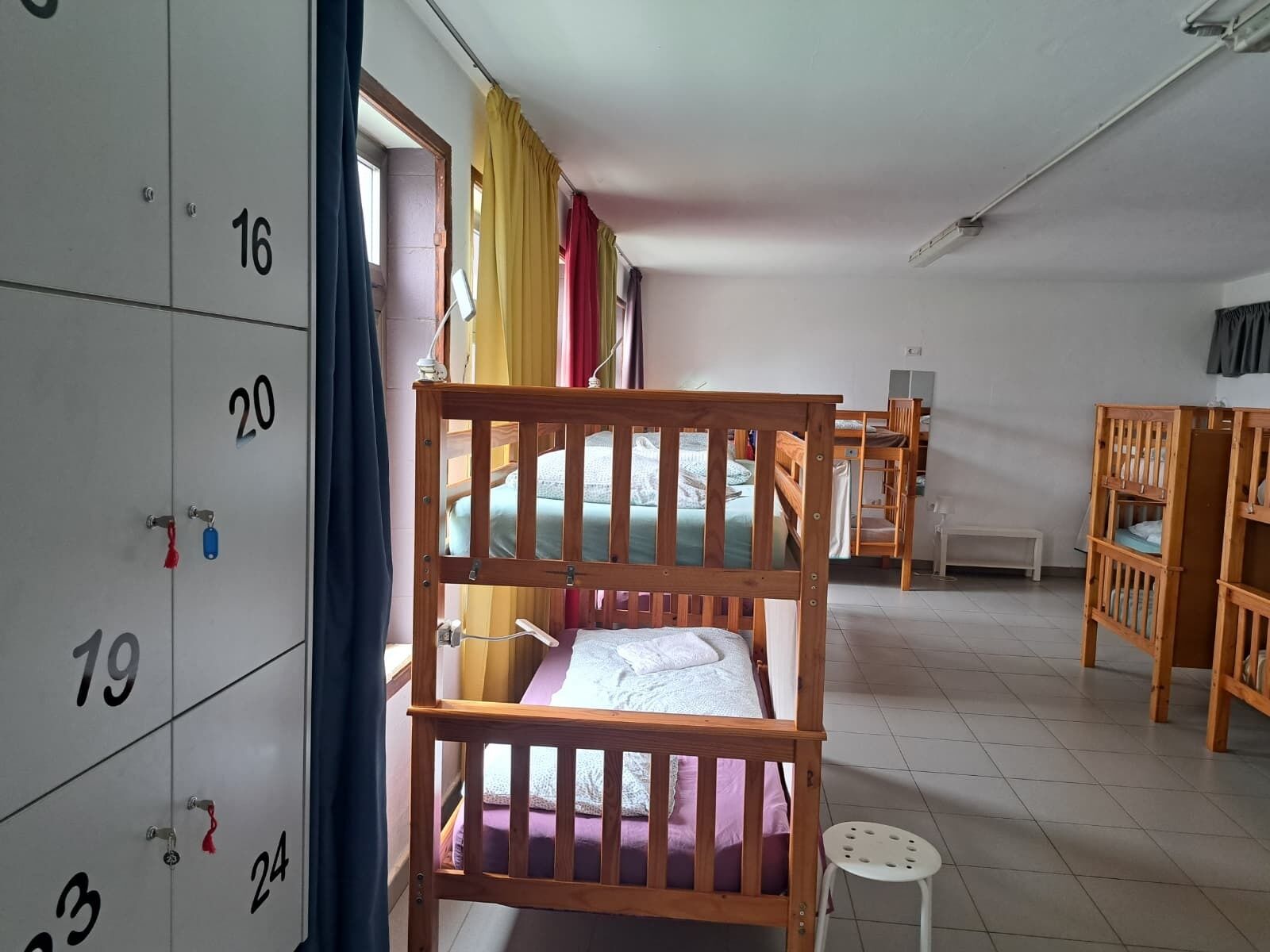A dormitory room with bunk beds, a desk, a chair, a small stool, and a wardrobe with numbered compartments.