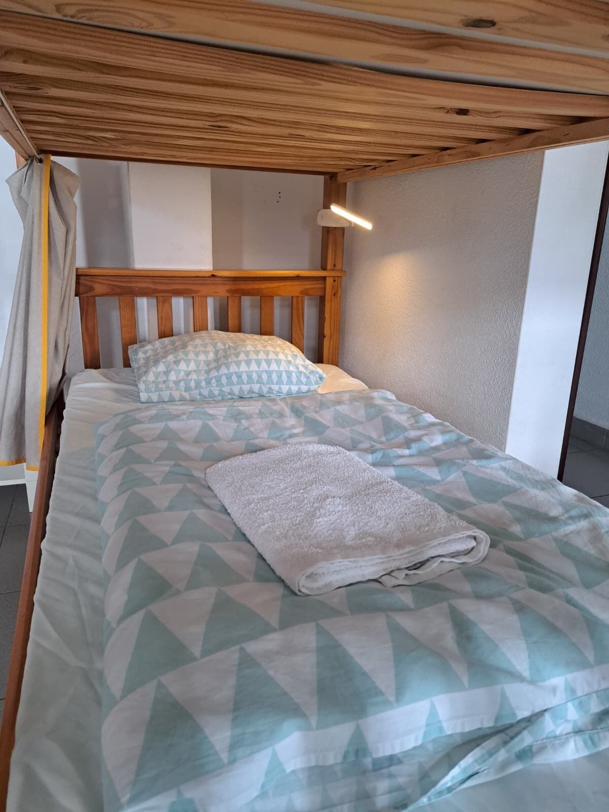 A single bed with a geometric patterned bedspread, a wooden headboard, and a white towel folded on the bed.