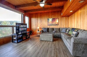 A wooden-paneled room with a sectional sofa, a television, and a window overlooking a forest. - Picturesque Cottage Retreat with a Private Hot Tub near Gatlinburg, Tennessee (Gatlinburg)