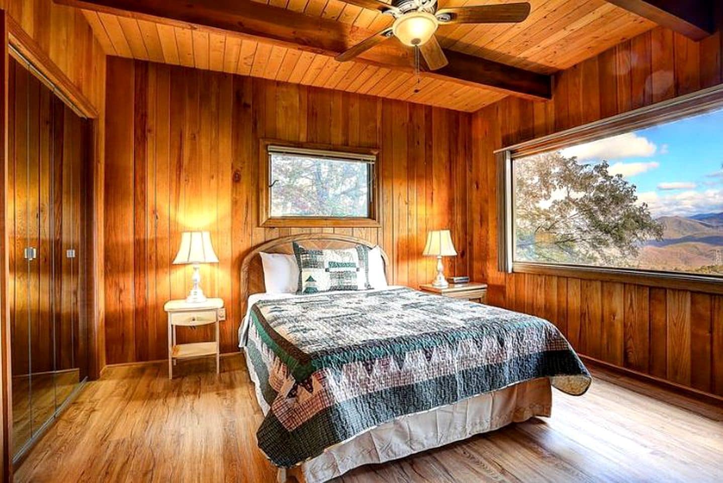 A wooden-paneled bedroom with a bed, two bedside lamps, a ceiling fan, and a large window with a mountain view.