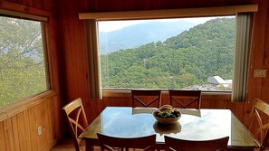 Ein Holzinnenraum mit einem Esstisch, Stühlen, einer Obstschale und einem großen Fenster mit Blick auf einen bewaldeten Berg.