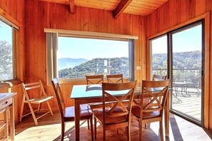 Interior em madeira com uma mesa de jantar e cadeiras, janelas amplas com vista para as montanhas e uma porta de correr para um deck externo.