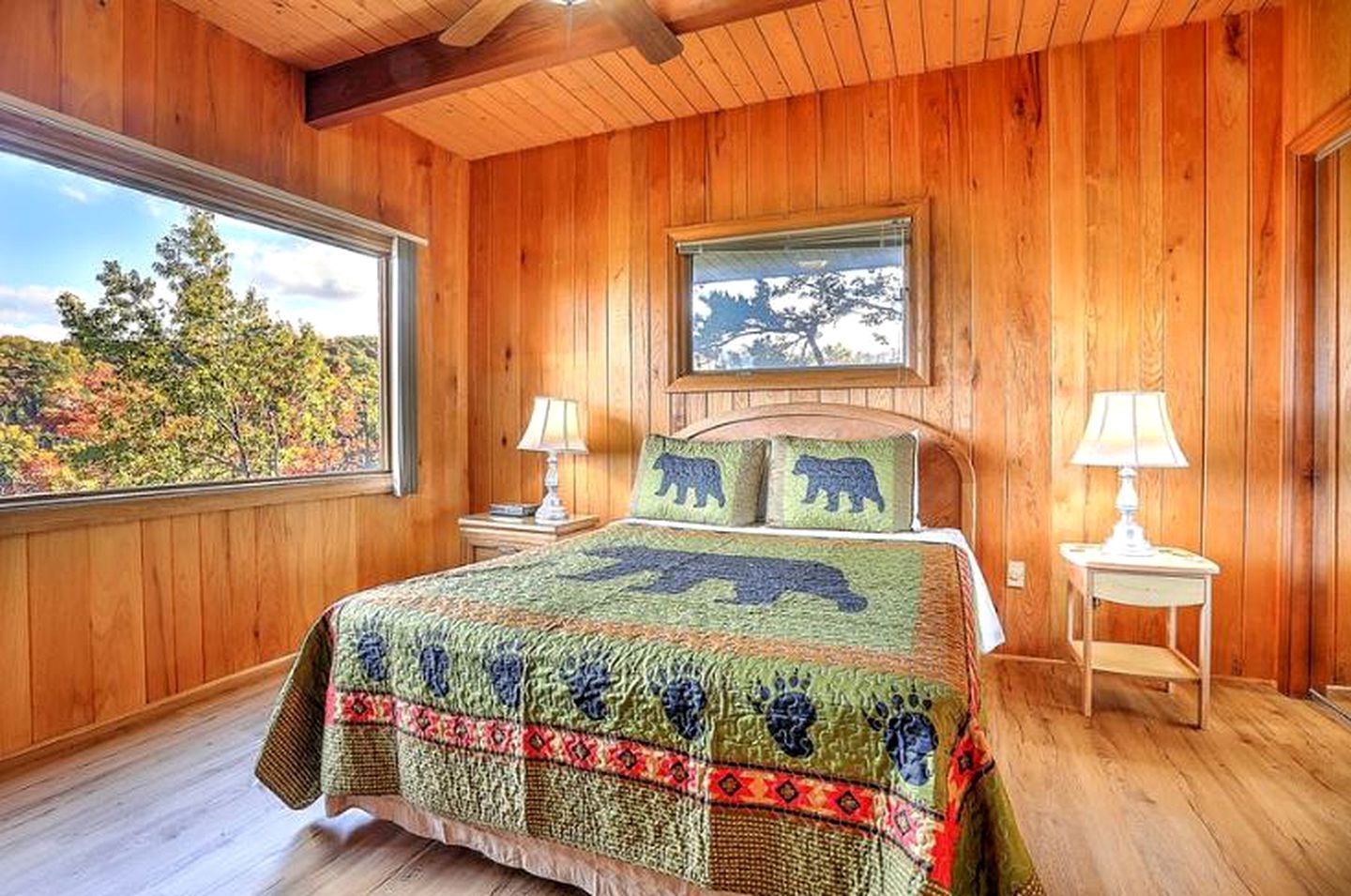A wooden-paneled bedroom with a large bed, two bedside tables, and a window with a view of trees.