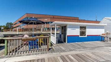 Un edificio frente a la playa con fachada azul y blanca, una terraza de madera y un área de estar cubierta con mesa y sillas.