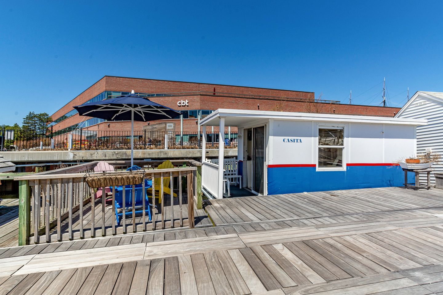 Un edificio frente a la playa con fachada azul y blanca, una terraza de madera y un área de estar cubierta con mesa y sillas.