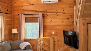 A wooden cabin interior with a sofa, a TV mounted on a wall, a window with curtains, and a ceiling fan.