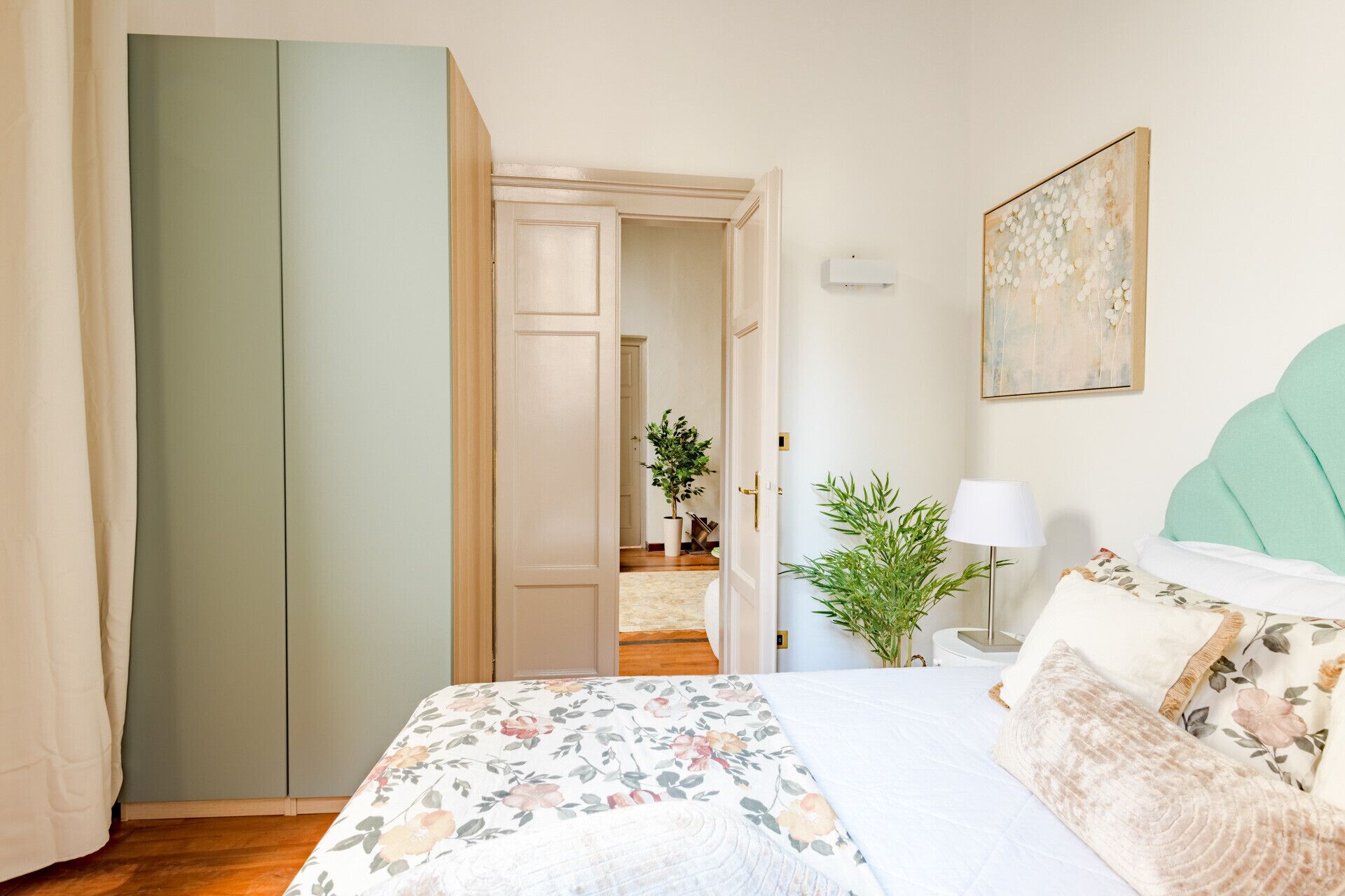 A bedroom with a bed, a nightstand, a lamp, a painting, and a door leading to another room.
