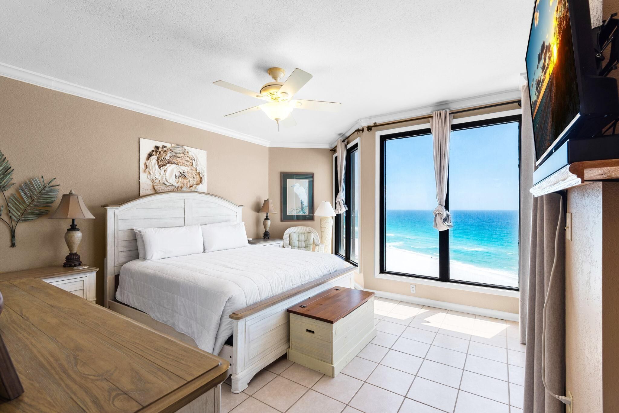 A bedroom with a bed, a ceiling fan, a TV, a window with a view of the ocean, and a wooden bench.