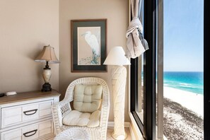 Ein Korbstuhl neben einem Fenster mit Blick auf den Strand, ein gerahmtes Vogelgemälde und eine Lampe auf einem Holztisch.