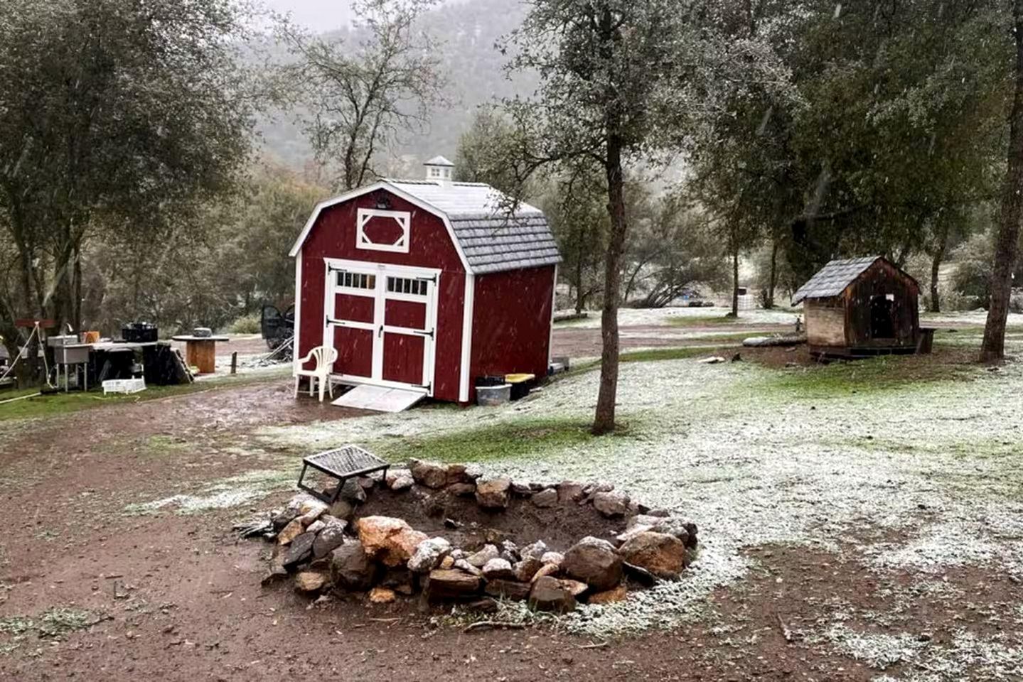 Een rode schuur met een witte deur en ramen, een klein tuinhuisje, en een vuurkorf van steen in een landelijke omgeving.