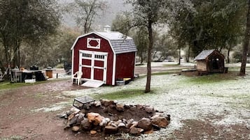 Een rode schuur met een witte deur en ramen, een klein tuinhuisje, en een vuurkorf van steen in een landelijke omgeving.
