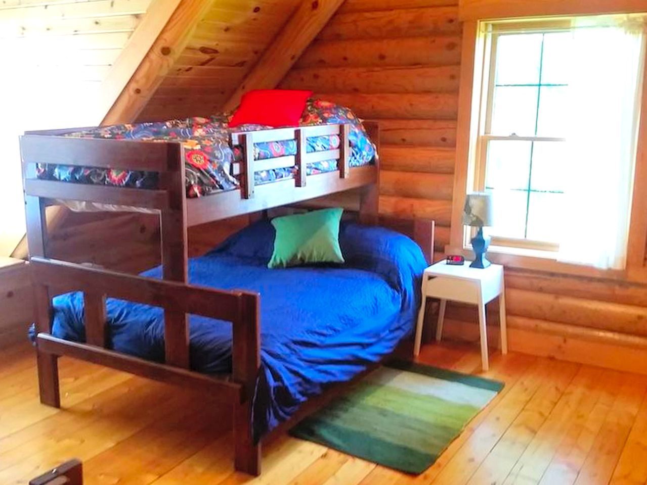 A bunk bed with a patterned top bunk and a blue bottom bunk in a log cabin room with wooden floors and a window.