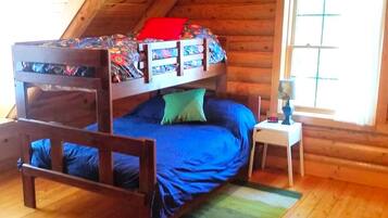 A bunk bed with a patterned top bunk and a blue bottom bunk in a log cabin room with wooden floors and a window.