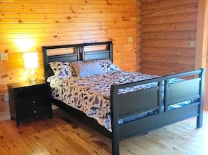 A wooden cabin bedroom with a bed, nightstand, and wall-mounted lamp.