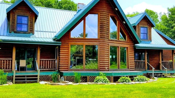 Une cabane en rondins dotée d’une véranda, de grandes fenêtres et d’un toit en métal.