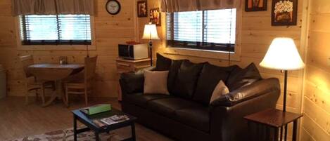A cozy log cabin interior with a leather sofa, a small table, a lamp, and a dining area.
