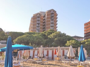 A beach with sun loungers and umbrellas, a tall building in the background, and trees. - Relaxing sea Front Apartment (Lignano Sabbiadoro)