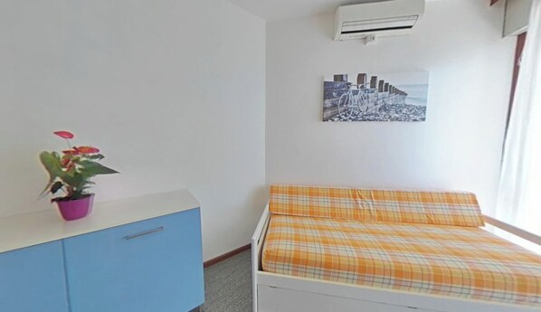 A bedroom with a bed, a dresser, a potted plant, an air conditioner, and a cityscape painting. - Relaxing sea Front Apartment (Lignano Sabbiadoro)