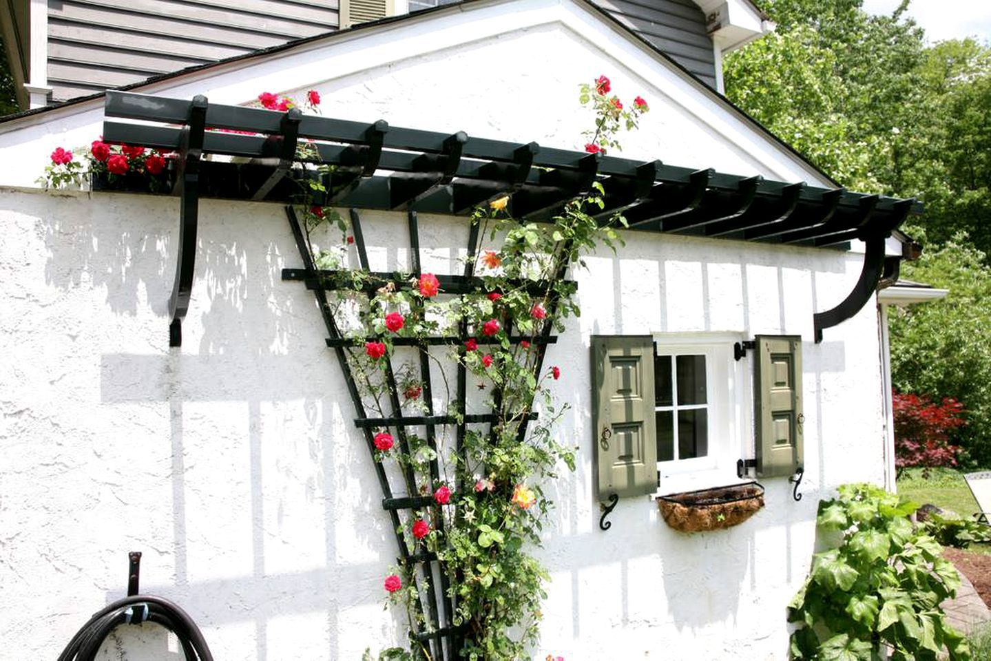 Un edificio blanco con una celosía negra trepando por una pared, flores rojas y una ventana con persianas verdes.