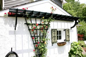 A white building with a black trellis climbing a wall, red flowers, and a window with green shutters.