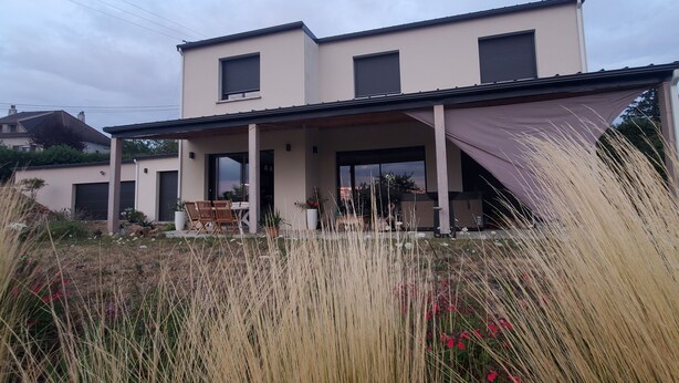 A modern two-story house with a covered patio, large windows, and a garden with tall grasses and red flowers.