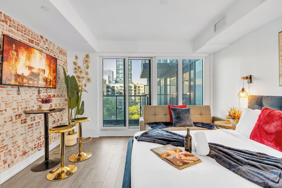 Une chambre d’hôtel moderne avec un grand lit, un canapé, un téléviseur fixé à un mur en briques et une vue sur la ville à travers de grandes fenêtres.